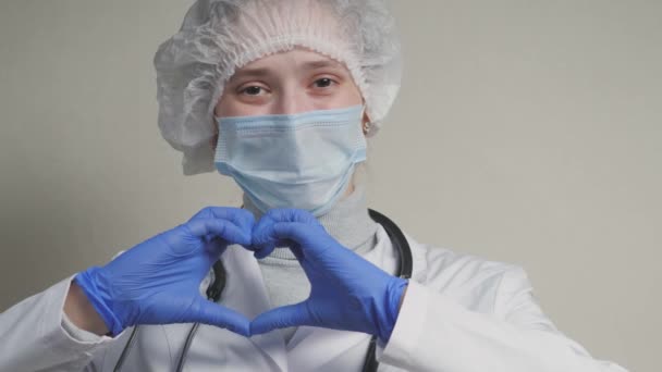 Un médico con una máscara azul y guantes sonríe y muestra su corazón, un cardiólogo profesional en una clínica de salud, una cita con un pediatra en el consultorio, una buena enfermera, pandemia de coronavirus — Vídeos de Stock