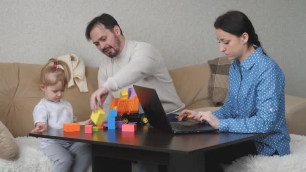 Mamá es una freelancer que trabaja en la computadora en casa, el niño y el papá están jugando con cubos coloridos, el desarrollo de habilidades motoras para niños, estilo de vida familiar, los padres están criando a una hija pequeña — Vídeos de Stock