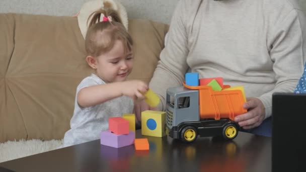 Батько грає зі своєю маленькою донькою в іграшкових кубиках і іграшковому авто, дитина весело проводить час в приміщенні з татом, кольорові заходи для дітей-аутистів, розвиток дитини з раннього віку, дідуся і — стокове відео