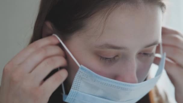 Mladá dívka si nasadí lékařskou masku, zatímco stojí u okna, žena zhluboka dýchá a usmívá se, dívá se do kamery, zdravotní a lékařské koncepce, detailní portrét, 4k — Stock video