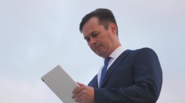 Mavi gökyüzüne karşı tableti olan takım elbiseli bir işadamı, işi ilerletiyor, modern teknolojilerle ilk oluyor, online uygulamalar geliştiriyor ve çalışma programları geliştiriyor.