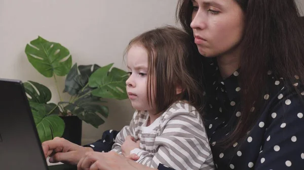 Женщина с маленьким ребенком работает за компьютером, фрилансер в домашнем офисе в ноутбуке с ребенком, деловая жизнь удаленно, смотрит на дисплей и печатает на клавиатуре, сидя на — стоковое фото