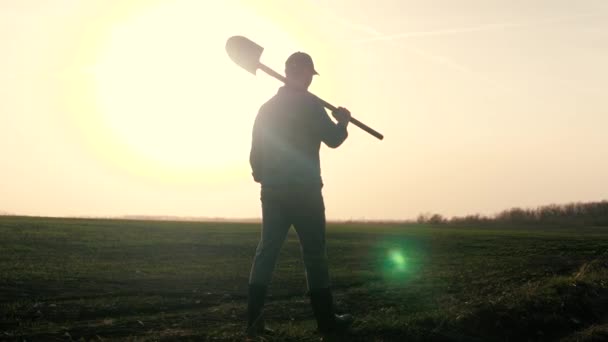 Agronom kráčí po černém oraném poli při západu slunce, farmář s lopatou v rukou, pracuje na poli pro dělníka, úsvit na obloze pro životní aktivity v zemědělství — Stock video