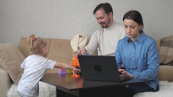 Mamá es una freelancer que trabaja en la computadora en casa, el niño y el papá están jugando con cubos coloridos, el desarrollo de habilidades motoras para niños, estilo de vida familiar, los padres están criando a una hija pequeña — Vídeos de Stock