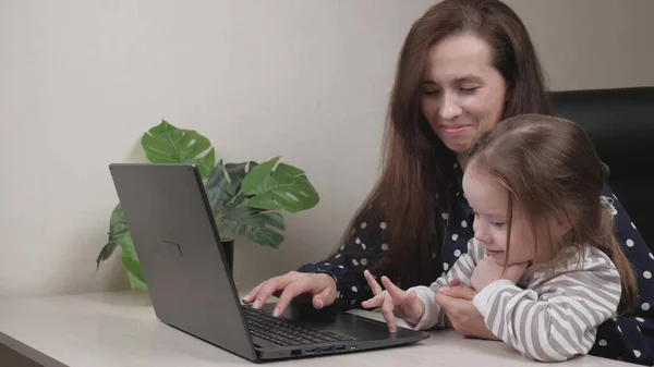 A freelance woman with a child in her arms sits at a table and prints an authors work on a laptop, a report for colleagues remotely. Kid interferes with a frustrated tired mother at the computer and — Stock Photo, Image