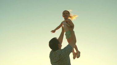 Mutlu baba güneşin parıltısıyla bir çocuğu gökyüzüne fırlatır, çocuk bir uçak gibi yükseğe uçar ve güler, küçük kız spa ile açık havada oynar, mutlu aile kavramı