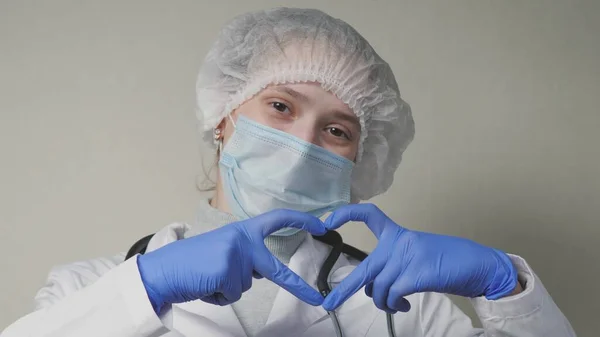 Врач в синей маске и перчатках улыбается и показывает свое сердце, профессионального кардиолога в поликлинике, встречу с педиатром в офисе, хорошую медсестру, пандемию коронавируса — стоковое фото