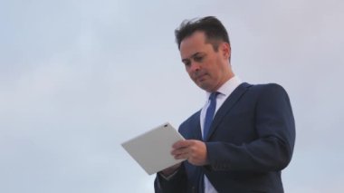 Takım elbiseli bir iş adamı, mavi gökyüzüne karşı bir tablette online sipariş verir, modern yetişkin hayatı, cihaz teknolojileri, bir uygulama, eğitim ve ileri eğitim alanında çalışır.