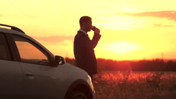 Un homme d'affaires en faux costume boit du café au coucher du soleil du soir dans le ciel debout à côté de la voiture, la liberté de voyager au volant, se concentrer et penser à l'avenir à l'extérieur, un homme à l'aube — Video