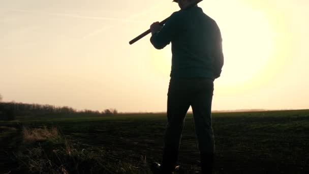 Een agronomist loopt door een zwart geploegd veld bij zonsondergang, boer met een schep in zijn handen, werk in het veld voor de arbeider, dageraad in de lucht voor het leven in de landbouw — Stockvideo