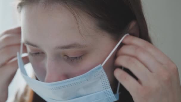 Jong meisje zet een medisch masker op terwijl ze bij het raam staat, het vrouwtje ademt diep en glimlacht, kijkend naar de camera, gezondheidszorg en medisch concept, close-up portret, 4k — Stockvideo