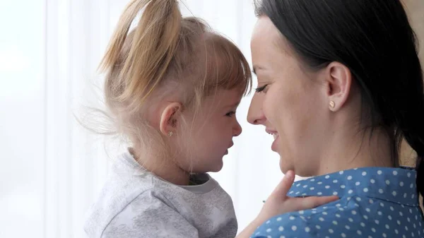 A mother holds her daughter in her arms while standing by the window and plays with her nose, happy family life, a girl with her mother on weekends indoors, an adult nanny looks after a small child — Stock Photo, Image