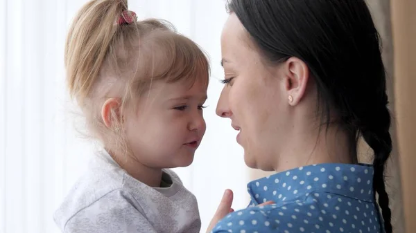 Uma mãe segura sua filha em seus braços enquanto está ao lado da janela e brinca com seu nariz, vida familiar feliz, uma menina com sua mãe nos fins de semana dentro de casa, uma babá adulta cuida de uma criança pequena — Fotografia de Stock