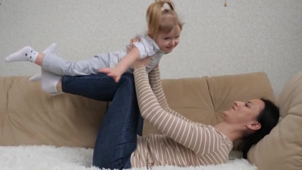 Счастливая маленькая девочка играет со своей матерью в самолете, лежа на диване, девушка парит в воздухе, лежа на коленях матери, женщина занимается фитнесом с ребенком на руках — стоковое видео