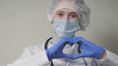 Mavi maskeli ve eldivenli bir doktor gülümser ve kalbini gösterir, bir sağlık kliniğinde profesyonel bir kardiyolog, ofisteki bir çocuk doktoru, iyi bir hemşire, koronavirüs salgını