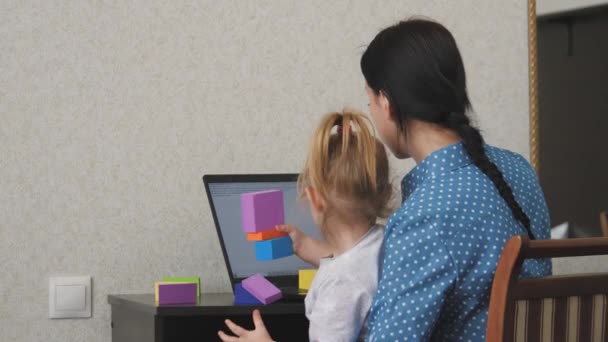 Mãe trabalha em um laptop com uma criança pequena em seus braços, a criança coleta uma torre de cubos coloridos, uma mulher freelance cuida do bebê, trabalhando em casa, sentada em casa, ganhando dinheiro online — Vídeo de Stock