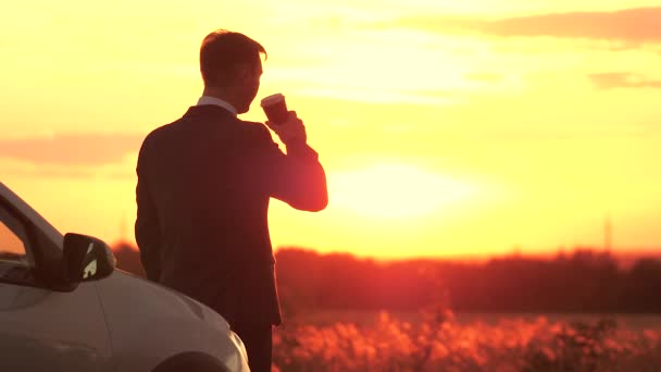 Een zakenman in een neppak drinkt 's avonds koffie bij zonsondergang in de lucht naast de auto, vrijheid van reizen achter het stuur, concentreren en denken aan de toekomst buiten, een man bij dageraad — Stockvideo