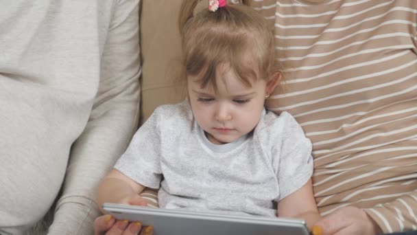 一个小孩和他的父母坐在沙发上玩平板电脑，一个小孩在小摆设中观看训练录像，一个婴儿在网上的应用，一个现代家庭的概念 — 图库视频影像