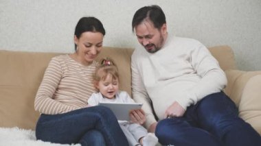 Küçük bir çocuk, annesi ve babasıyla kanepede oturuyor ve tablet oynuyor, çocuk bir alet ekranında eğitim videosu izliyor, bebek için internet uygulamaları, modern aile kavramı.