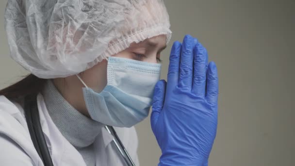 Un médico reza, un trabajador médico con una bata blanca, guantes azules, una gorra y una máscara pide ayuda a Dios, una enfermera está molesta en el trabajo en un hospital, un médico se preocupa por la salud de los pacientes, la fatiga y — Vídeos de Stock