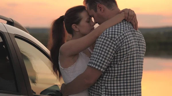 Un hombre y una mujer se abrazan en la orilla de la playa mientras están de pie al atardecer junto al coche, una pareja casada viajando en transporte personal, conociendo el amanecer juntos, relación en un par de —  Fotos de Stock