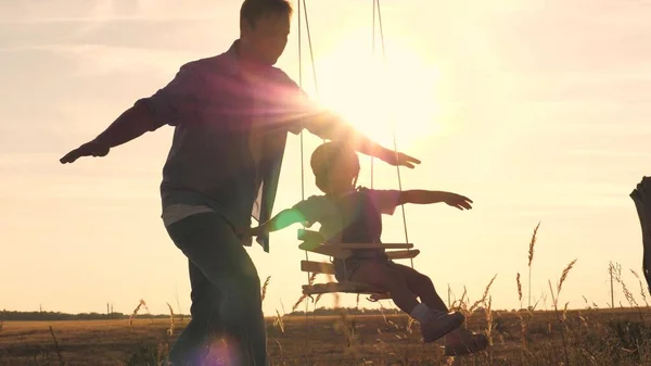 Happy little child with a teddy bear and dad is riding a swing at sunset in the park, playing the plane flying in the air. Daughter and father are having fun at dawn. Happy family life concept. Kid — Stock Photo, Image