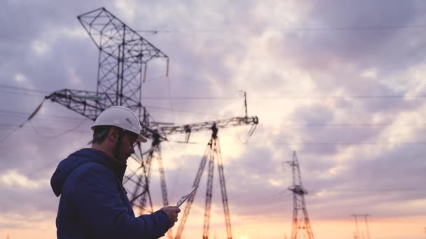 Silueta de ingeniero de pie en el campo con torres eléctricas. Ingeniero eléctrico con pilón eléctrico de alto voltaje al atardecer. Trabajadores del poder en el trabajo concepto — Vídeo de stock