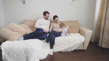 Annesi ve babasıyla birlikte küçük bir kız, bir tablet çalıyor, kapalı bir alanda aile eğlencesi, insanlar için modern teknolojiler, online bir mağazada online alışveriş, anne ve babasından bir çocuk