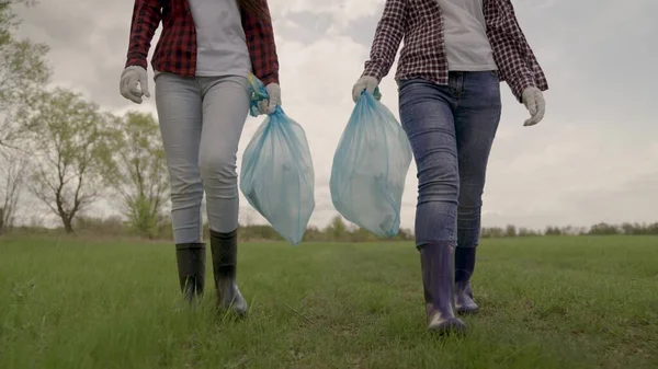 Les filles marchent sur un champ vert dans des bottes en caoutchouc et tiennent des sacs à ordures dans leurs mains, l'écologie de la propreté, la sensibilisation des bénévoles, la pollution, l'environnement, la famille heureuse, déchets en plastique, — Photo