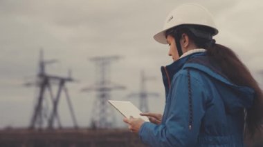 Yüksek gerilim santralinde çalışan bir mühendis gün batımında bir tablette çalışır, elektrik enerjisi tasarımı, endüstri konsepti, modern iş teknolojileri, bir elektrikçi bir cihaz üzerine bir proje okur.