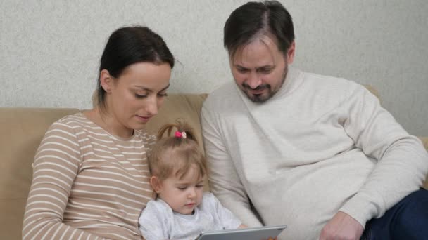 Un niño pequeño está sentado en el sofá con su madre y su padre y jugando una tableta, niño viendo un video de entrenamiento en una pantalla de gadget, aplicaciones de Internet para el bebé, el concepto de familia moderna — Vídeo de stock