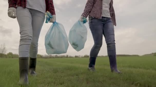 Meninas caminham em um campo verde em botas de borracha e seguram sacos de lixo em suas mãos, ecologia da limpeza, conscientização voluntária, poluição, meio ambiente, família feliz, lixo plástico, — Vídeo de Stock