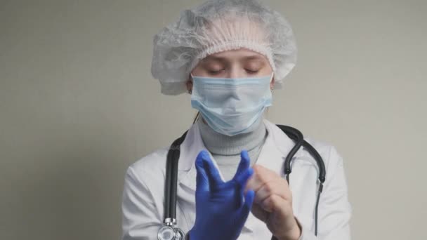 En læge i en beskyttende maske, et stetoskop og en hætte lægger på medicinske handsker på sine hænder, undersøger patienter, beskytter mod coronavirus, en epidemisk pandemi – Stock-video