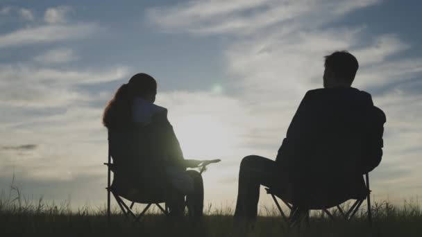 Sylwetka mężczyzny i kobiety widząc zachód słońca razem na niebie, ręka w rękę, podróże rodzinne w przyrodzie, koncepcja szczęśliwego życia, siedzenie na krzesłach i relaksujące spotkanie świtu, weekend — Wideo stockowe