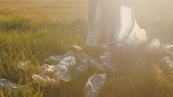 Eine Frau mit Handschuhen sammelt bei Sonnenuntergang Müll in einem Müllsack und bewahrt so eine saubere Ökologie von Luft und Erde, der Umwelt, der Erhaltung der Erde, Plastikmüll im grellen Licht der Sonne — Stockvideo