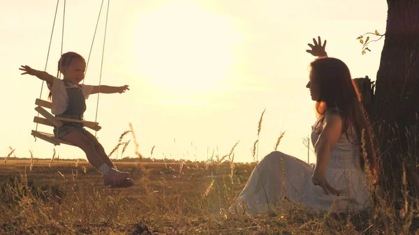 Een volwassen vrouw rolt een kind op een schommel in de stralen van de zonsondergang in de lucht, het concept van een gelukkig gezin, het kind en de moeder spelen buiten in het vliegtuig, zweven in de lucht en dromen om te vliegen — Stockfoto