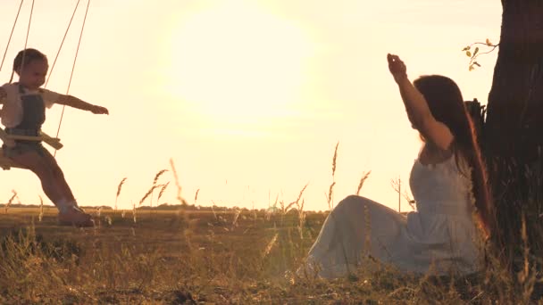 Een volwassen vrouw rolt een kind op een schommel in de stralen van de zonsondergang in de lucht, het concept van een gelukkig gezin, het kind en de moeder spelen buiten in het vliegtuig, zweven in de lucht en dromen om te vliegen — Stockvideo