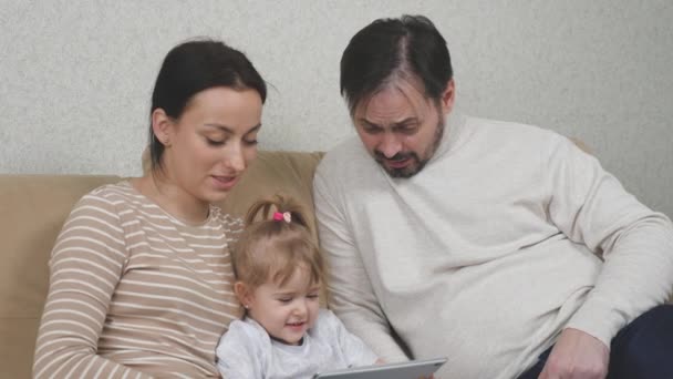 Dziecko i rodzice siedzą na kanapie z tabletem w rękach i śmieją się, online ucząc dziecko w aplikacji za pomocą Internetu, weekend zabawy w domu, szczęśliwa koncepcja rodziny — Wideo stockowe
