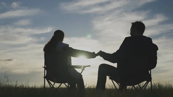 Silhouette of a man and a woman seeing off the sunset together in the sky, hand in hand, family travel in nature, the concept of a happy life, sitting on chairs and relaxing meeting the dawn, weekend — Stock Photo, Image