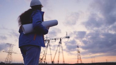 Elektrik santrallerinin arka planına karşı gün batımında çalışan bir elektrikçi, elektrik şebekeleri ve kablolarının bakımı için çalışan genç bir çalışan mühendisi, voltaj temin etmek için bir proje üzerinde çalışıyor.