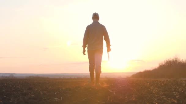 Rolnik chodzi z tabliczką w ręku na ziemi o zachodzie słońca na niebie, pracujący agronomista zajmuje się uprawą ziemi na polu, nadszedł czas, aby zapłodnić ziemię dla sadzonek — Wideo stockowe