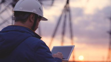 Bir elektrikçi, elektrik kulelerinin arka planına karşı tablette çalışır, bir mühendis elektrik akımının seviyesini ölçer, bir elektrik şebekesi de ışık kaynağı sağlar.