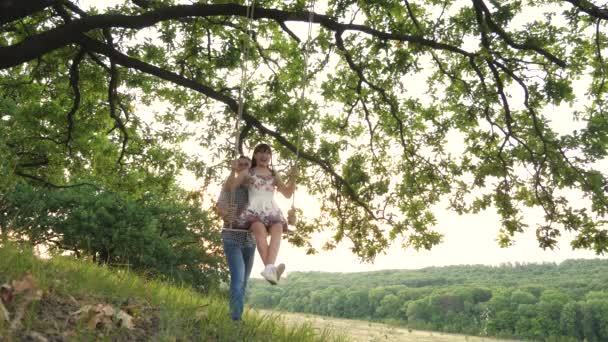 父亲在秋千上摇曳着他的成年女儿，笑着说："一个快乐的家庭，爸爸和一个女孩在大自然里玩得开心，在空中飞舞，在夏天玩得开心极了。 — 图库视频影像
