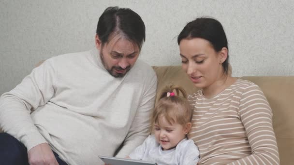 Dziecko i rodzice siedzą na kanapie z tabletem w rękach i śmieją się, online ucząc dziecko w aplikacji za pomocą Internetu, weekend zabawy w domu, szczęśliwa koncepcja rodziny — Wideo stockowe