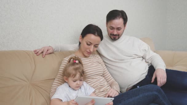 Een klein kind zit op de bank met zijn moeder en vader en speelt een tablet, kind kijkt naar een trainingsvideo in een gadget display, internet toepassingen voor baby, het concept van moderne familie — Stockvideo