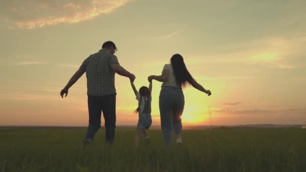 Sebuah keluarga yang bahagia berjalan di lapangan dengan seorang anak kecil saat matahari terbenam di langit, berjalan ibu, ayah dan anak melompat riang di lapangan di malam hari, tim bepergian dengan gembira bermain — Stok Video