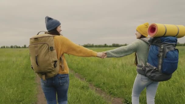 Dos chicas hipster con mochilas van en una caminata cogidas de la mano, felices viajeros milenarios están caminando a lo largo de un camino de tierra y en busca de aventura, tiempo para soñar en la naturaleza, personas activas — Vídeos de Stock