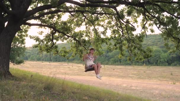 Een volwassen meisje zwaaiend op een schommel vastgebonden aan een boom in de natuur, zwevend in de lucht in de wind en lachend, het concept van een gelukkig leven — Stockvideo