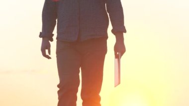 Çizmeli bir çiftçi gün batımında elinde tabletle yolda yürüyor, tarımda modern aletlerin kullanılması, bir arazi çiftliğinde çalışmak, bahçede tarım işi, sebze bahçesi mevsimi