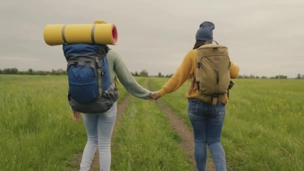 Twee hipster meisjes met rugzakken gaan op een wandeling hand in hand, gelukkige duizendjarige reizigers lopen langs een onverharde weg en op zoek naar avontuur, tijd om te dromen in de natuur, actieve mensen — Stockvideo